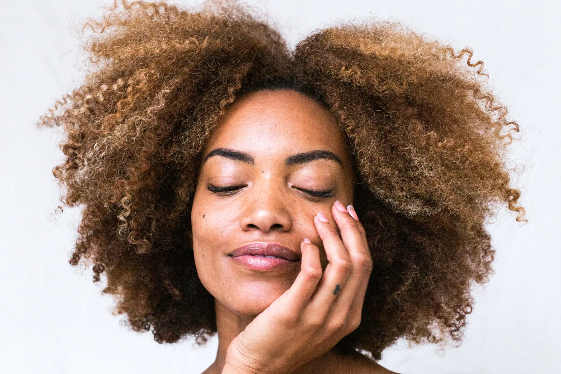 A woman with curly hair, eyes closed, resting her face in her hand